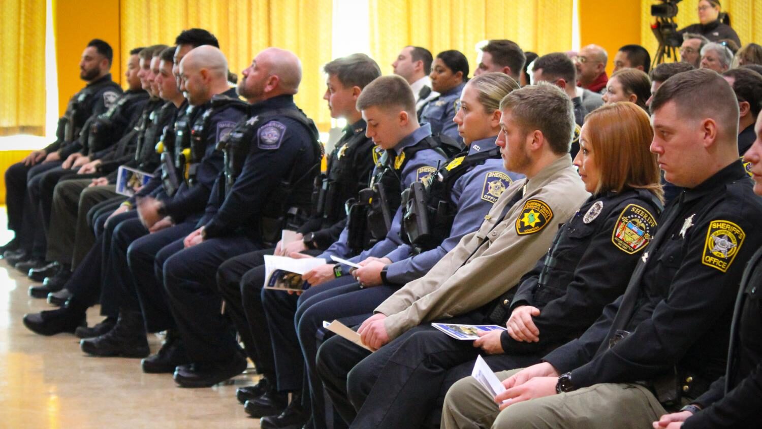 Sheriffs in uniform sit in folding chairs at a graduation ceremony